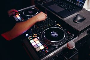 A close-up view of a DJ's hands expertly manipulating a mixing console, flanked by a laptop and microphone, underscoring the contemporary evolution of music and entertainment within the broader history of nightlife.